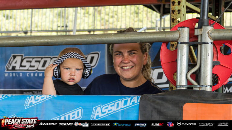 Cloe Phend smiles beside Dax, who wears checkered hearing protection, behind the trackside railing at the 2026 Georgia Peach State Classic. A candid moment from the race weekend’s Good Times photo gallery.