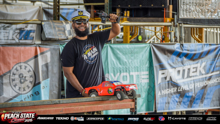 A smiling RC racer in a captain’s hat raises a transmitter beside an orange short course truck at the 2026 Georgia Peach State Classic. A candid moment from the race weekend’s Good Times photo gallery.